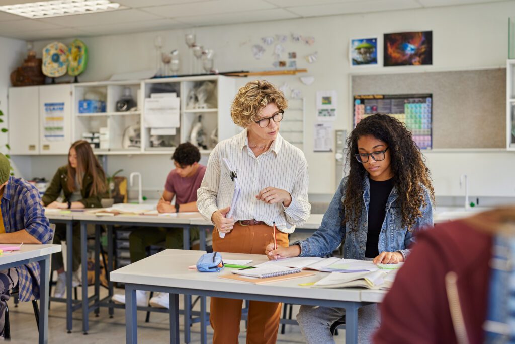 Teacher guiding female student while classmates are studying in background.